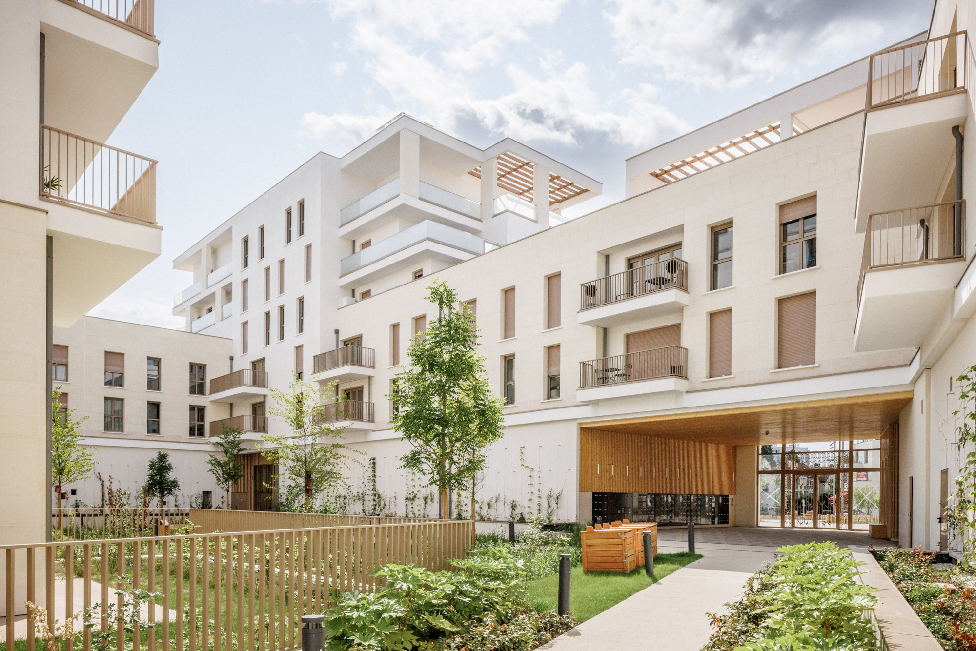 Logements collectifs ZAC Jean Zay à Antony (Hauts-de-Seine), conçus par Cussac Architectes et Associés (ca+a), avec façades blanches en pierre, balcons et jardin paysager autour d’un porche en bois.